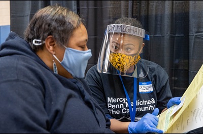 Meghan Lane-Fall, MD, MSHP, of Anesthesiology, wears a mask and a face shield labeled “Dr. Meghan” while speaking to a seated woman wearing a blue mask.